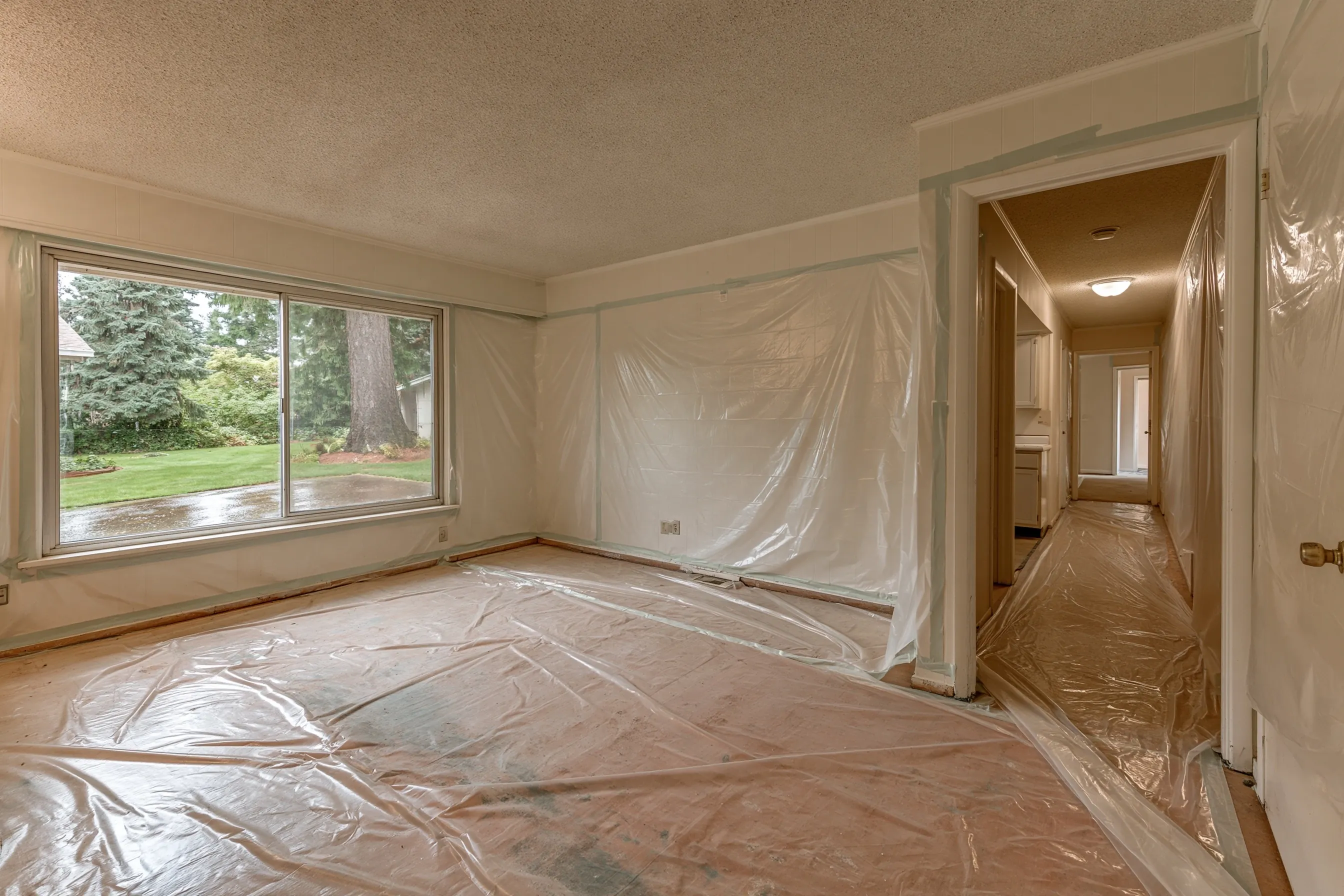 An empty living room and hall with a textured ceiling, plastic sheeting over the floor and part of the walls, and a large window onto a lawn