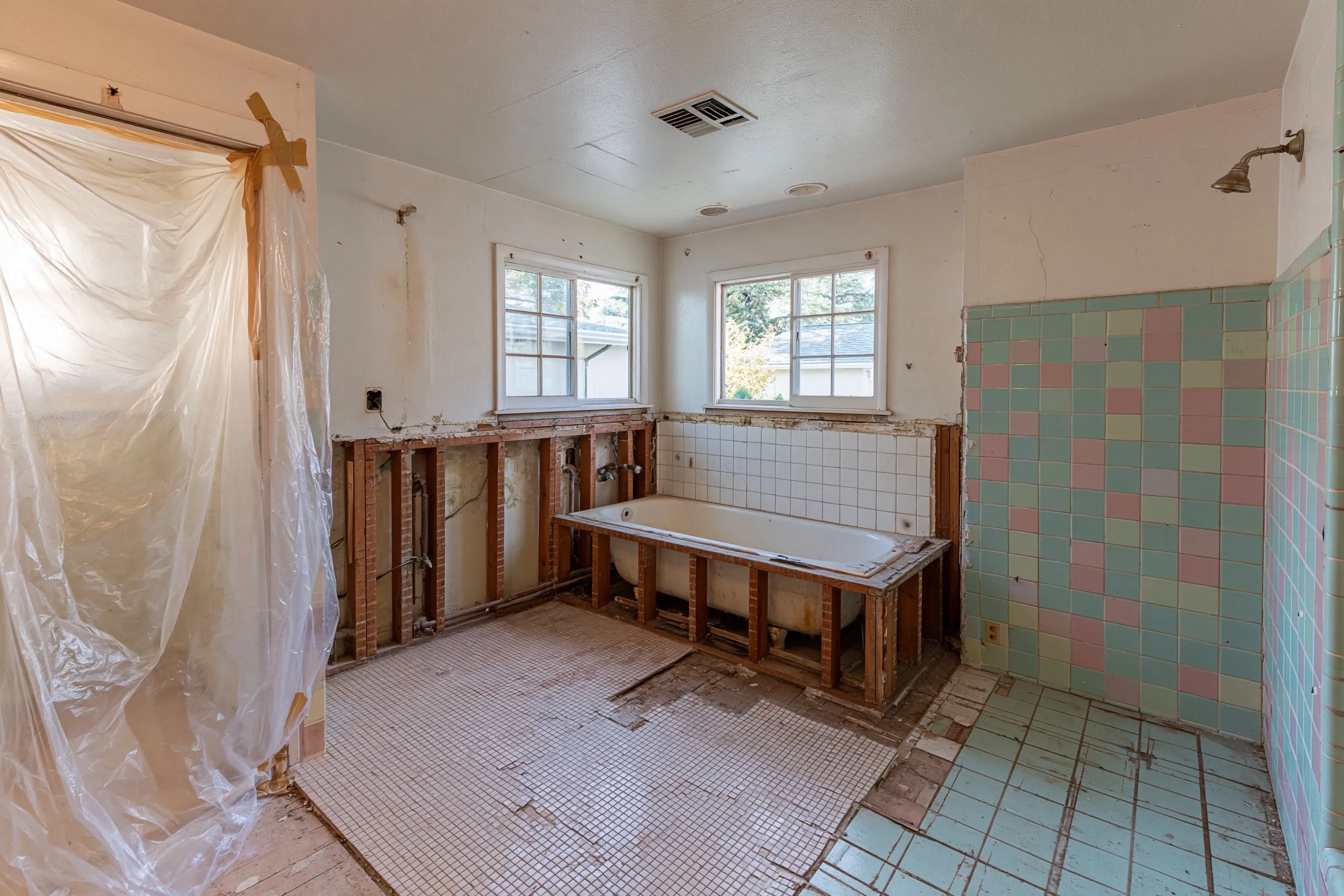 A bathroom opened for a remodel, with the wall around the tub removed to the studs, small floor tiles lifted in places and plastic hung at one side